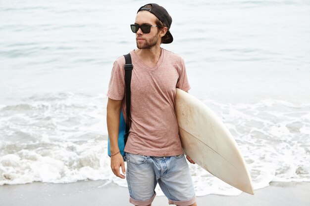 Young man wearing fashionable sunglasses and snapback holding surfboard in his hand