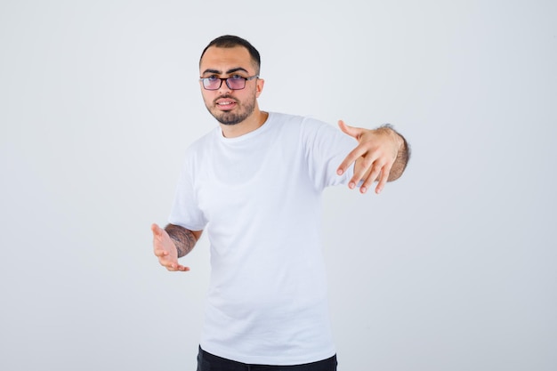 Young man wearing glasses and stretching hands as holding something in white t-shirt and black pants and looking happy