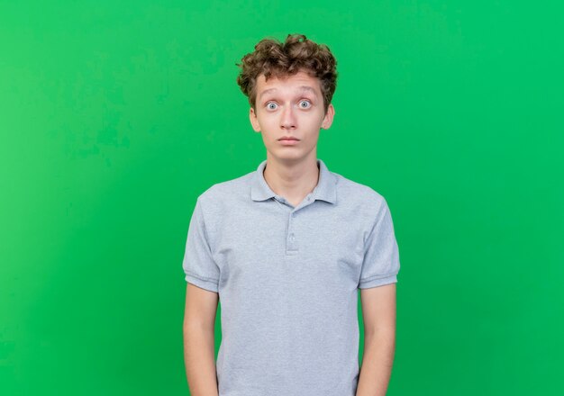 Young man wearing grey polo shirt  surprised and amazed with wide opened eyes standing over green wall