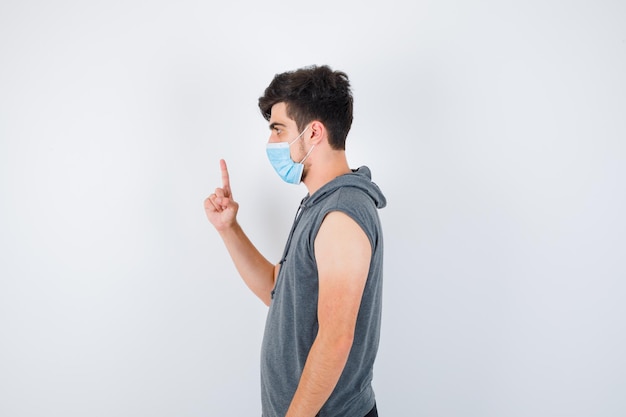 Young man wearing mask while raising index finger in gray t-shirt and looking serious