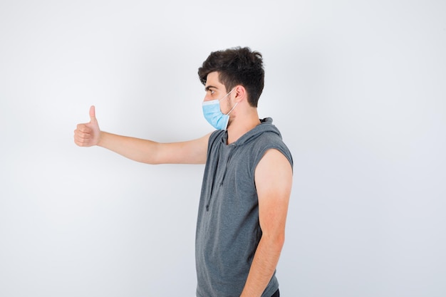 Young man wearing mask while showing thumb up in gray t-shirt and looking serious