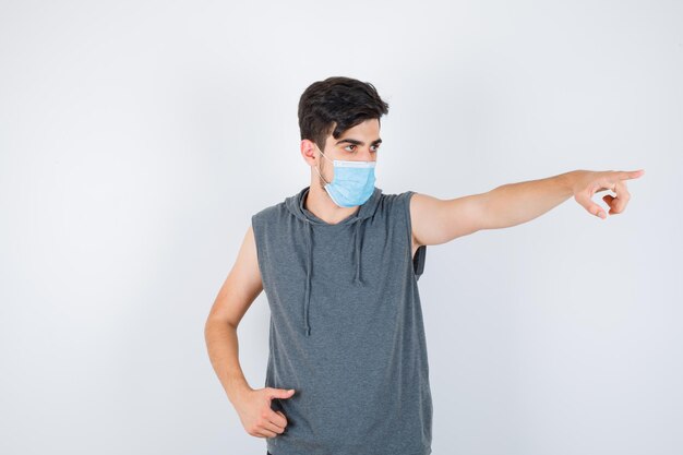 Young man wearing mask while showing thumb up and pointing right in gray t-shirt and looking serious