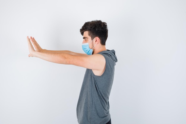 Young man wearing mask while stretching hands toward left in gray t-shirt and looking serious