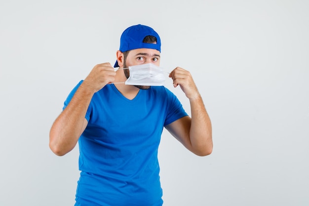 Young man wearing medical mask in blue t-shirt and cap and looking careful