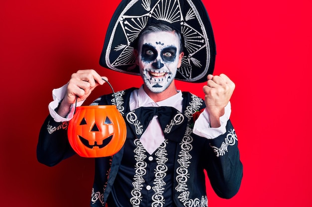 Young man wearing mexican day of the dead costume holding pumpkin screaming proud celebrating victory and success very excited with raised arms