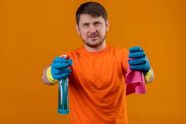 Бесплатное фото Молодой человек в оранжевой футболке и резиновых перчатках держит чистящий спрей и ковер, уверенно улыбается