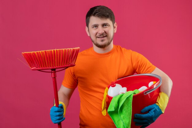 청소 도구와 청소 도구와 걸레 오렌지 티셔츠를 입고 젊은 남자 분홍색 배경 위에 서 청소 준비가 카메라 긍정적이고 행복 준비가 웃고