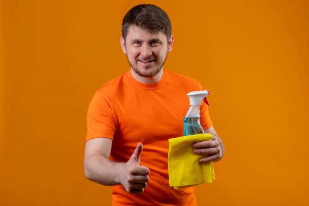 Free photo young man wearing orange t-shirt holding cleaning spray and rug smiling cheerfully positive and happy looking at camera showing thumbs up standing over orange background