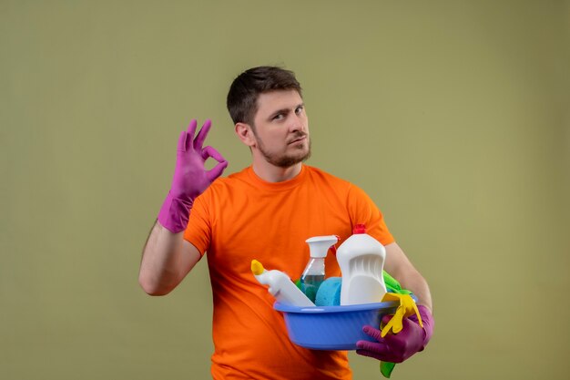 Young man wearing orange t-shirt and rubber gloves holding basin with cleaning tolls looking confident doing ok sign standing over green backgroun