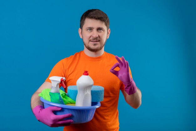 Young man wearing orange t-shirt and rubber gloves holding basin with cleaning tools