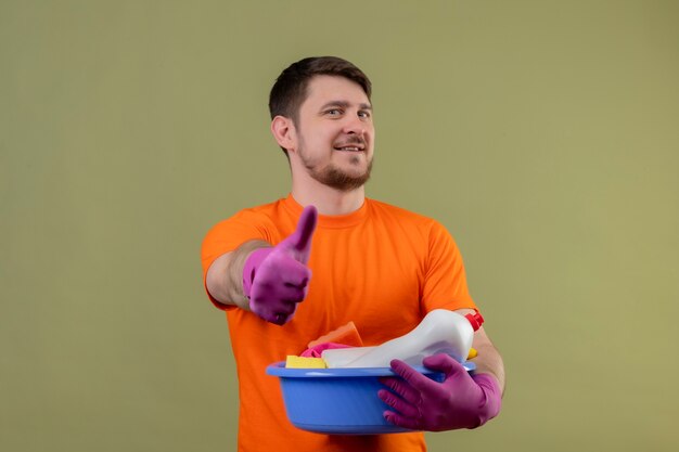 Young man wearing orange t-shirt and rubber gloves holding basin with cleaning tools