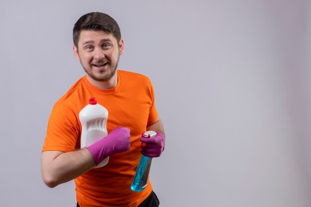 Молодой человек в оранжевой футболке и резиновых перчатках держит бутылки с чистящими средствами и весело улыбается