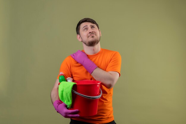 Young man wearing orange t-shirt and rubber gloves holding bucket with cleaning tools looking tired and overworked touching shoulder having pain standing over green wall