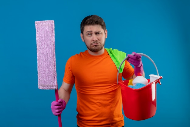 Young man wearing orange t-shirt and rubber gloves holding bucket with cleaning tools and mop