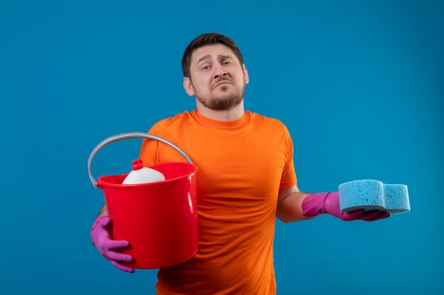 Молодой человек в оранжевой футболке и резиновых перчатках держит ведро с чистящими средствами и губкой