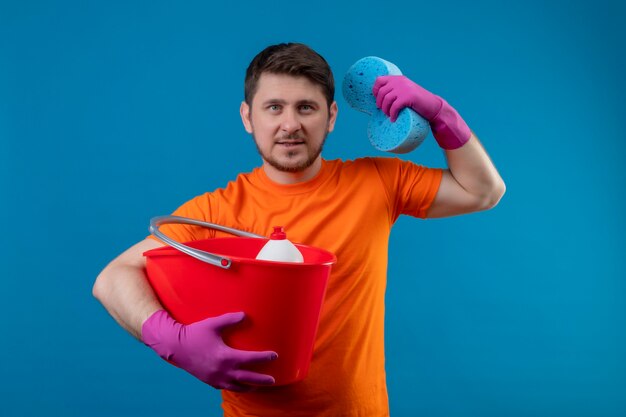 Молодой человек в оранжевой футболке и резиновых перчатках держит ведро с чистящими средствами и губкой