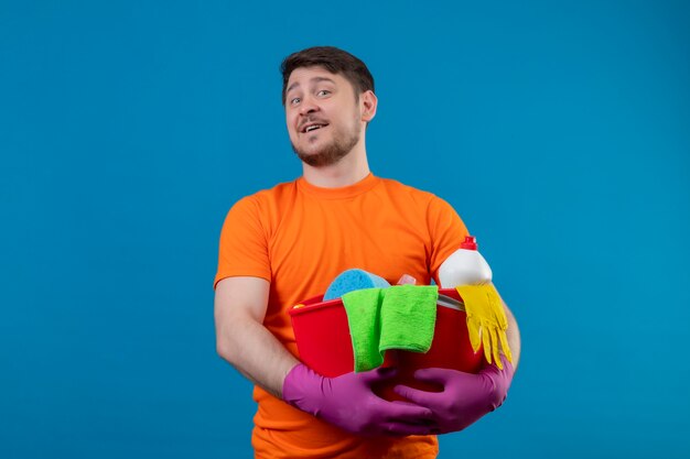 Молодой человек в оранжевой футболке и резиновых перчатках держит ведро с чистящими средствами