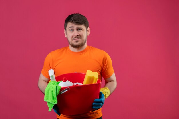 Young man wearing orange t-shirt and rubber gloves holding bucket with cleaning tools