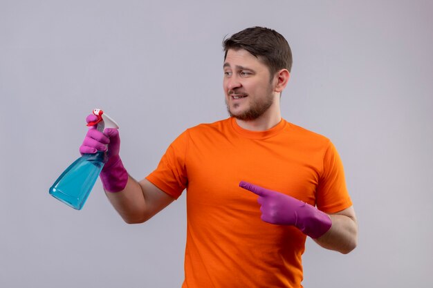 Young man wearing orange t-shirt and rubber gloves holding cleaning spray looking at it pointing with finger