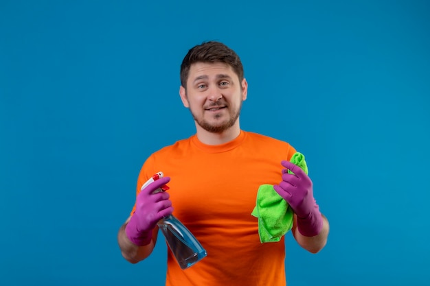 Free photo young man wearing orange t-shirt and rubber gloves holding cleaning spray and rug