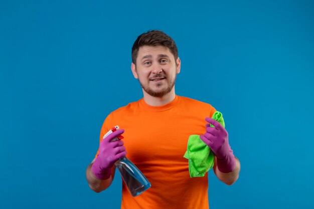 Young man wearing orange t-shirt and rubber gloves holding cleaning spray and rug