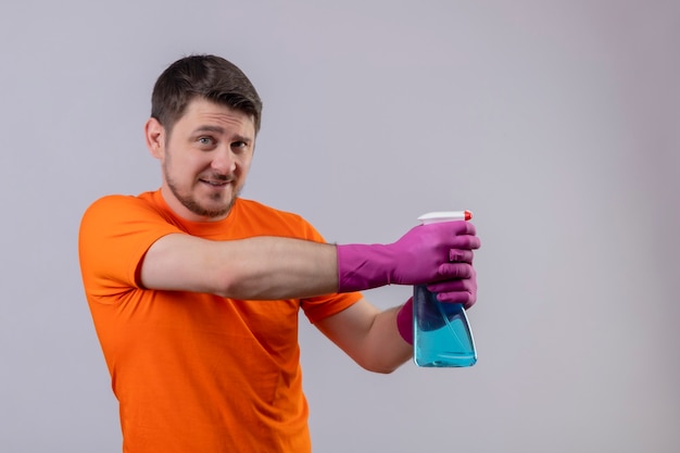 Giovane uomo che indossa la maglietta arancione e guanti di gomma che tengono spray per la pulizia sorridente utilizzando spray in piedi sul muro bianco