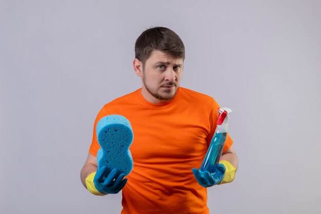 Young man wearing orange t-shirt and rubber gloves holding cleaning spray and sponge with skeptic expression on face standing over white wall