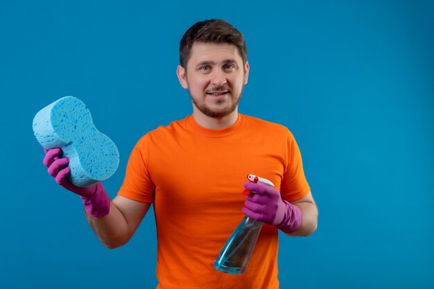 オレンジ色のtシャツとクリーニングスプレーとスポンジを保持しているゴム手袋を身に着けている若い男