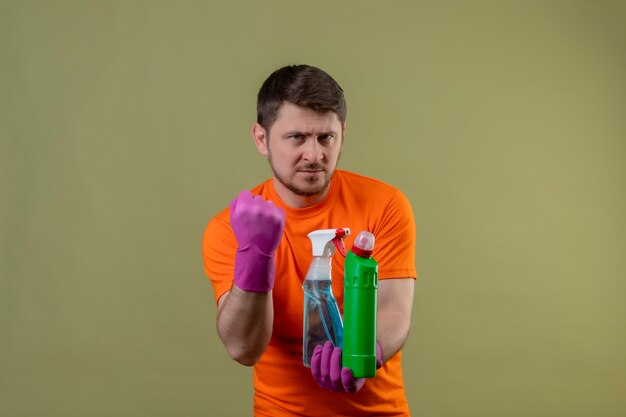 Young man wearing orange t-shirt and rubber gloves holding cleaning supplies looking confident clenching fist with serious expression on face ready to clean concept standing over green wall