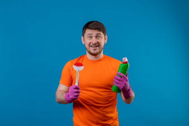 Young man wearing orange t-shirt and rubber gloves holding cleaning supplies and scrubbing brush