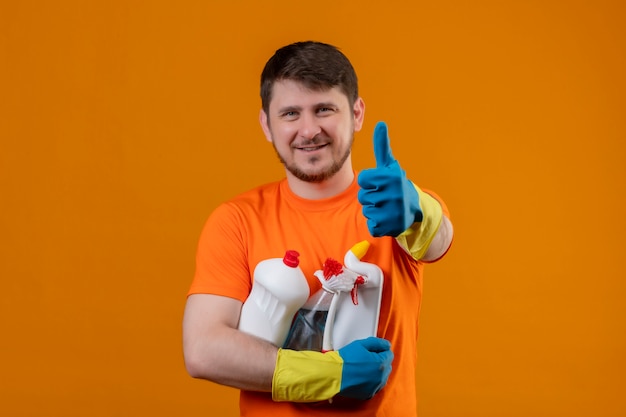 Молодой человек в оранжевой футболке и резиновых перчатках держит чистящие средства, весело улыбаясь и радостно глядя в камеру, показывая пальцы вверх, готовые очистить концепцию на оранжевом фоне