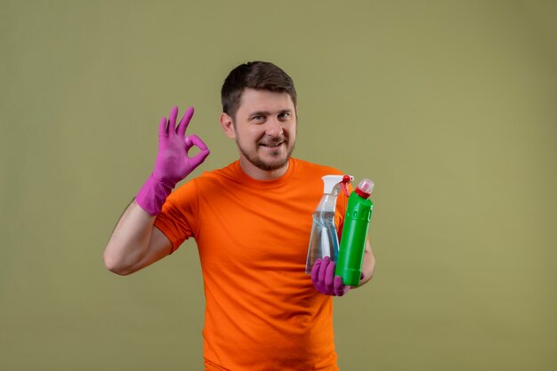 Young man wearing orange t-shirt and rubber gloves holding cleaning supplies smiling happy and positive doing ok sign standing over green wall