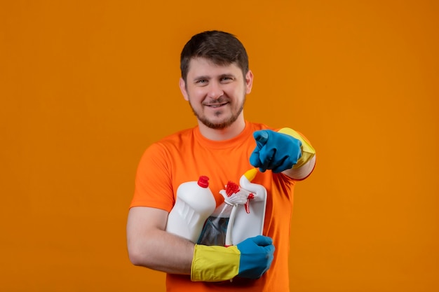 Young man wearing orange t-shirt and rubber gloves holding cleaning supplies smiling positive