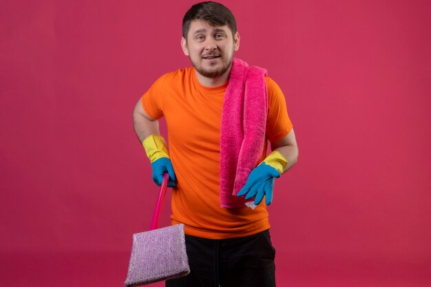 Young man wearing orange t-shirt and rubber gloves holding mop with raised arm as asking a question standing over pink wall