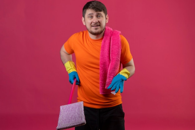 オレンジ色のtシャツとピンクの壁の上に立って質問をすることを調達の腕でモップを保持しているゴム手袋を着用して若い男