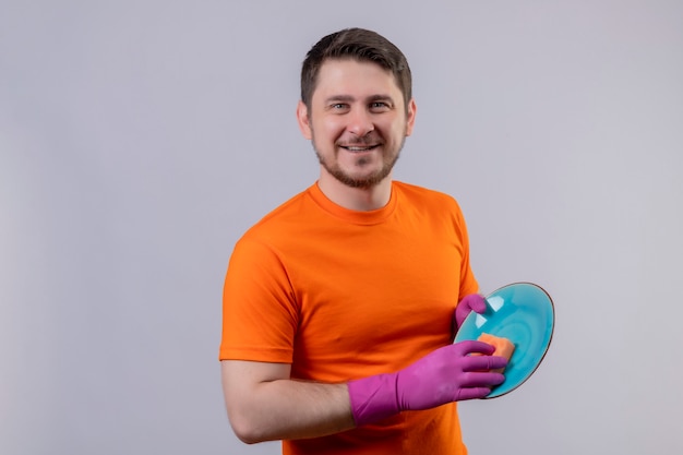 Young man wearing orange t-shirt and rubber gloves washing plate looking at camera smiling positive and happy standing over white background