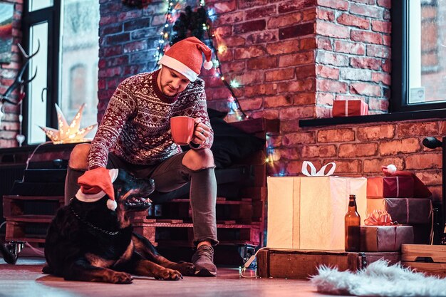 装飾されたリビングルームで彼のかわいい犬と一緒にクリスマスの時期にコーヒーとカップを保持しているサンタの帽子をかぶっている若い男。