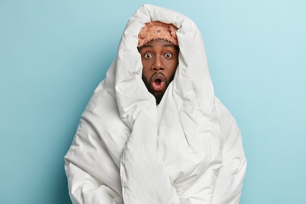 Young man wearing sleep mask and wrapped in white blanket