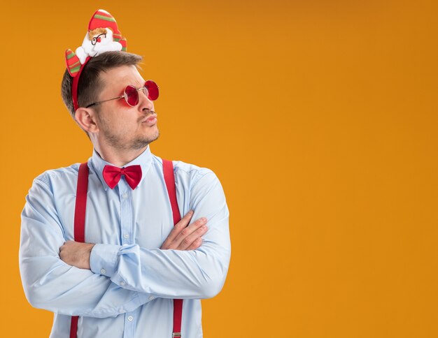 Young man wearing suspenders bow tie in rim with santa and red glasses looking aside with serious face standing over orange background