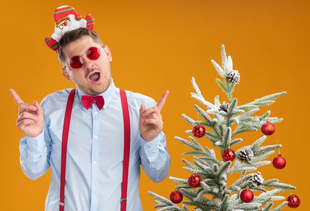 サスペンダーの蝶ネクタイを身に着けている若い男は、サンタと赤い眼鏡がクリスマスツリーの隣に立って幸せで陽気なオレンジ色の壁に人差し指を示しています