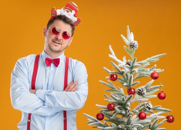 Young man wearing suspenders bow tie in rim with santa and red glasses standing next to christmas tree looking at camera with confident expression with arms crossed over orange background