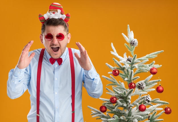 오렌지 벽에 공황 상태에서 외치는 크리스마스 트리 옆에 서있는 산타와 빨간 안경 테두리에 멜빵 나비 넥타이를 착용하는 젊은 남자