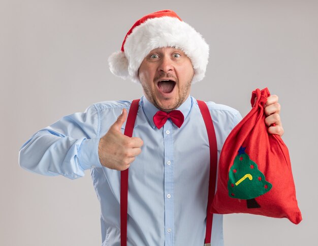 Young man wearing suspenders bow tie in santa hat holding santa claus bag full of gifts looking at camera happy and excited showing thumbs up standing over white background