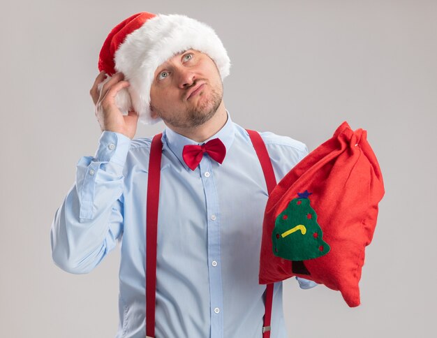 Young man wearing suspenders bow tie in santa hat holding santa claus bag full of gifts looking up puzzled standing over white background