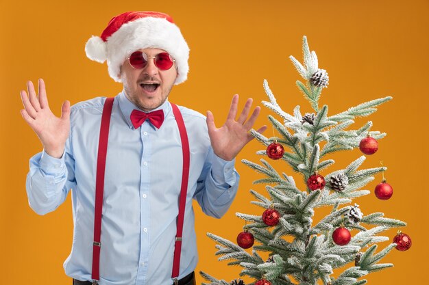 サスペンダーを身に着けている若い男は、オレンジ色の壁の上のクリスマスツリーの近くに立っている腕を上げて幸せで驚いたサンタ帽子と赤い眼鏡の蝶ネクタイ