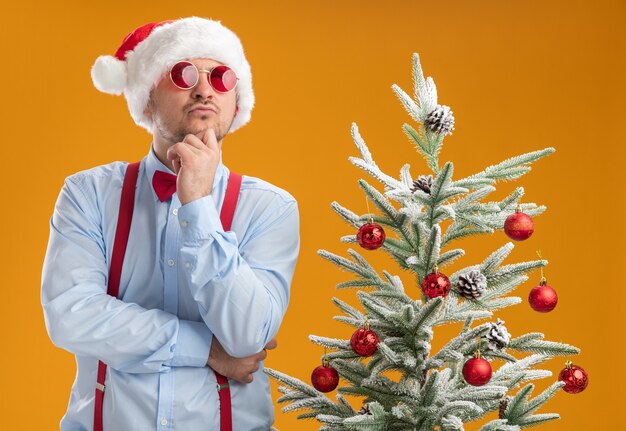 Молодой человек с подтяжками и галстуком-бабочкой в новогодней шапке и красных очках смотрит озадаченно, стоя возле елки над оранжевой стеной