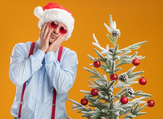 Young man wearing suspenders bow tie in santa hat and red glasses standing next to christmas tree looking at camera confused and very anxious over orange background