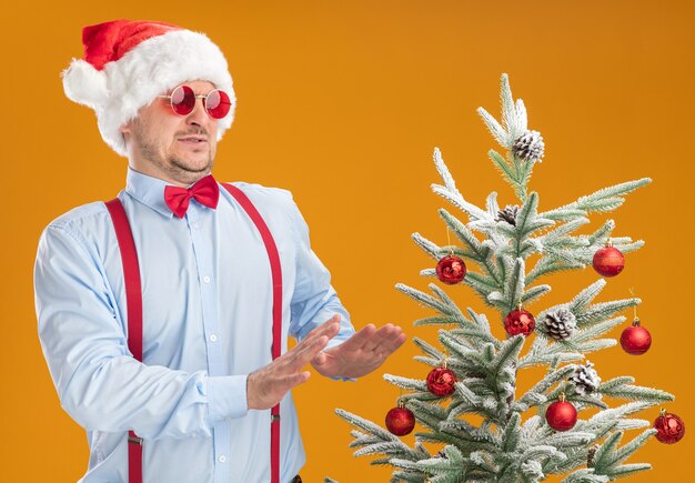 Young man wearing suspenders bow tie in santa hat and red glasses standing next to christmas tree looking at it with disgusted expression holding hands out over orange wall