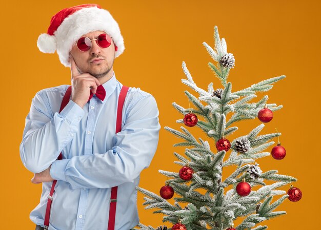 オレンジ色の壁を考えて顔に物思いにふける表現でクリスマスツリーの横に立っているサンタの帽子と赤い眼鏡でサスペンダー蝶ネクタイを身に着けている若い男