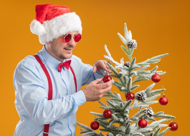 Young man wearing suspenders bow tie in santa hat and red glasses standing near christmas tree hanging toys on the tree happy and cgeerful over orange wall